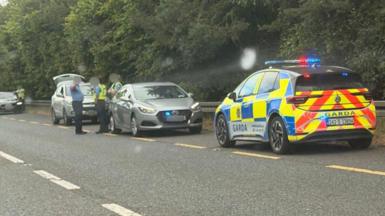 Garda officers and garda cars surround a small car on the roadside of the N2 near Carrickmacross. The officers are all wearing blue uniforms, but two are wearing yellow high-viz vests over their shirts. The boot of a hatchback vehicle behind them is open. 
