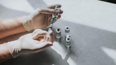 A gloved medical professionals' hand uses a syringe to extract liquid from some vials on a table. 