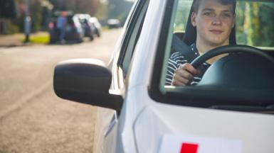 A male driver wearing a striped top sits in the driving seat of a car, with an L plate half visible. 