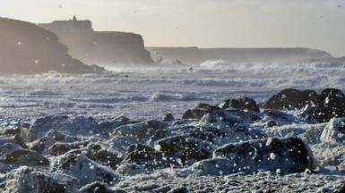 Rocks in the sea covered in foam as waves are whipped up by strong winds with stormy sea and coastline in background