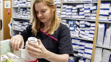 A female pharmacist with blonde hair and a black uniform works in the stock room of a pharmacist