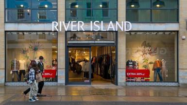 A group of young people walk past a River Island store with sale signs in the window