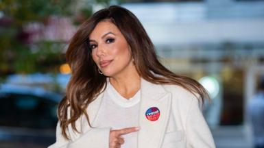 Eva Longoria, who has long brown hair and is wearing hoop earrings, a white trench coat and a plain white T-shirt, points with her right index finger to an 'I Voted' sticker over her left lapel