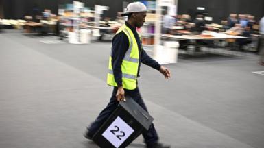 A man in a hi-vis and baseball cap worn backwards carries a ballot box with the number 22 on it. The background in the hall is out of focus.