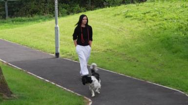 Kirsten Coutts, a woman with long black hair, is wearing white trousers and a black jumper rolled up at the arms as she walks along a tarmac path surrounded by grass. Her dog, a black and white spaniel, walks in front of her and she is wearing his lead around her neck.