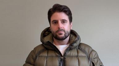 Alex Grossomanides has brown hair, brown eyes and a beard. He is wearing an olive green puffer coat and looking at the camera while standing in front of a white wall indoors.