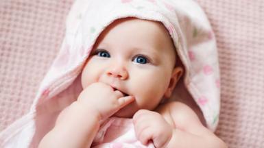 A baby sucks its fingers as it lies on a pink blanket. It is wrapped in a pale, floral towel, as if it has just had a bath.
