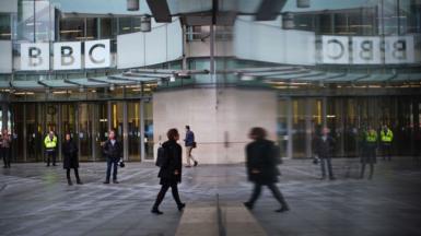 A person wearing a black jacket and carrying a backpack walks towards the entrance doors at BBC Broadcasting House in London on Monday, with the BBC logo in view.