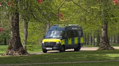 A yellow and blue police van is parked on a path among trees in Kensington Gardens