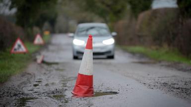 A red and white cone sits in a deep pothole. A car can be seen approaching in the background.