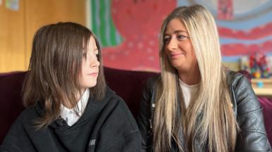 Hannah and Ashly are sitting in a classroom looking at each other. Ashly, the mother, has long blonde hair and is wearing a grey leather jacket and white blouse. She is smiling at her daughter who is looking into the distance. Hannah has shoulder-length dark hair and is wearing a white school shirt without a tie and black hoodie. 