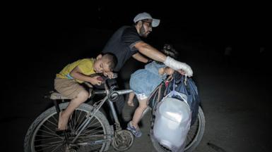 A man with a bandaged hand steers the handlebars of a bicycle while two small children lie on the front bars and seat. Some bags hang from the bike's handlebars as they journey in the night, with a bright light shining on to them.