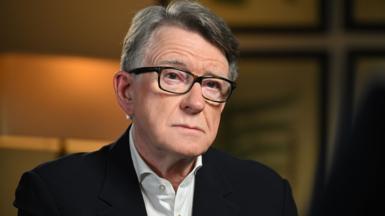 Lord Mandelson in front of a dark orange background looking serious