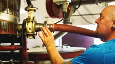 A man is operating beer brewing machinery. His hair is shaven and he is wearing a blue top with his hand on a metal valve.