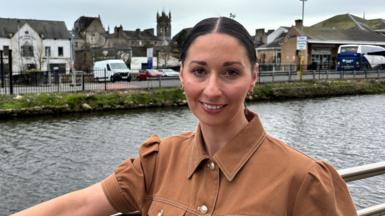 Rachel Duffy stands in front of Newry Canal. She is wearing a tan coloured shirt and has her dark hair pulled back from her face,
She is smiling