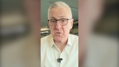 Dom Hughes looks straight at the camera. He wears glasses and a white shirt.