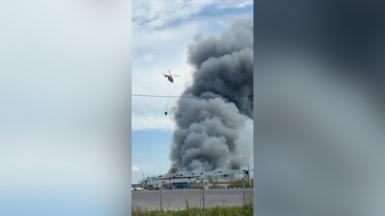 A helicopter carrying water flies towards a building with a huge plume of smoke erupting.