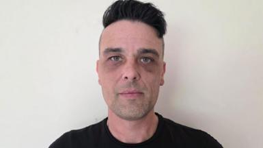 Kevin Brewer has black hair. He's wearing a black top and standing against a white wall.