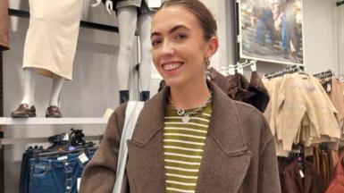 A woman with her hair tied up, in a yellow striped top and brown coat, smiles at the camera as she stands inside a Primark store