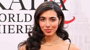 Izzy Escobar wears a black strappy formal dress with black gloves on the red carpet of the Devil Wears Prada 2 premiere. She wears a silver necklace and circular earrings 