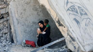 A Palestinian woman and a child rest under the rubble of a destroyed house in Khan Younis, in the southern Gaza Strip (16 October 2024)