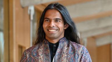 A person wearing a patterned jacket, with long flowing dark hair smiles as they walk towards the camera in the Scottish Parliament building
