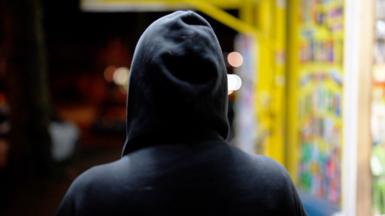 A person in a dark hoodie stands with their back to the camera, facing a brightly lit shop window filled with colourful items. The background is blurred with warm lights, creating a contrast between the dark figure and the vibrant display.
