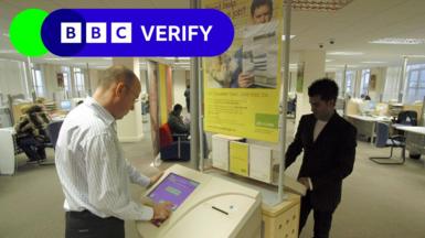 Interior of a Jobcentre showing a man standing at a Jobpoint computer terminal. The BBC Verify logo is in the top corner.