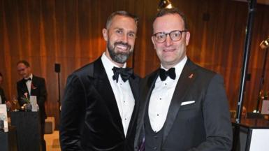 Jens Spahn (R) in glasses, a grey jacket and white shirt and bowtie beside his husband Daniel Funke who has as beard and a black velvet jacket with a white shirt and bowtie