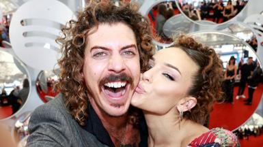 A man with curly brown hair, smiling widely at the camera, while a woman kisses him on the cheek