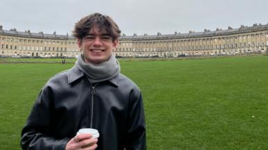 Sam, smiling into the camera and holding a takeaway coffee. His black leather jacket is zipped up and he is standing on the grass in front of the iconic Royal Crescent in Bath.