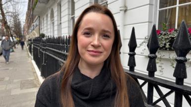 25-year-old Sophie stands outside an office on the pavement in front of black railings with a smile on her face.