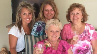 Anita LeBrun, 82, wears a bright pink floral top and corsage, smiling as she holds up a glass of white wine as female members of her family stand around her