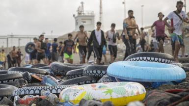 Rubber flotation rings sit on a beach in front of a line of arriving migrants in Ceuta.