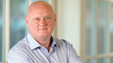 Campbell Scott staring straight at the camera. He is bald. He has blue eyes. He is wearing a light blue shirt with dark-coloured checks on it. He has his arms folded and is standing in front of a blurred background.