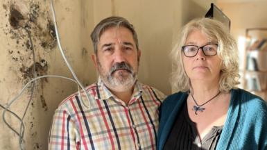 Tony and Bec Wadley standing next to a wall covered in black mould with electrical wires and sockets hanging from it