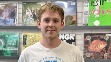 James, 25, with blonde hair wearing a cream T-shirt is smiling at the camera in front of row of records