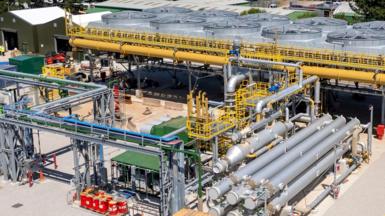 The United Downs power plant in Cornwall is shown, there are various silver pipes surrounding a central plant that has a series of large fan type structures surrounded by yellow pipes. There are trees in the background and the sky is blue.