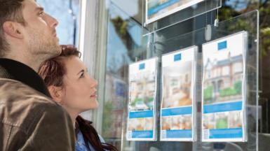 Close up of a young couple looking into an estate agents window showing both faces from the side  and information on three houses in the window 