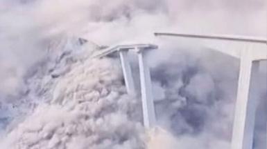 Bridge collapses leaving clouds of white smoke and dust, part of the remaining bridge is on the right of the image while the collapsed part of the bridge is on the left of frame.