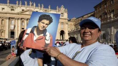 A woman in a crowd at the Vatican, wearing a white T-shirt and a pale blue baseball cap holds up a portrait picture of curly-haired Carlo Acutis, wearing a red jumper with blue collar, with the white handles of a backpack showing