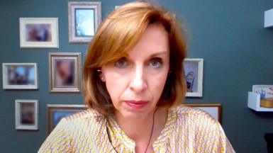 Dr Louise Newson looking at the camera - she has a jaw-length bob with a long fringe, which is a red/gold colour. She is wearing a patterned silk blouse and is wearing pink lipstick. She is wearing earphones as the shot was taken from a Zoom call. Behind her is a blue-painted wall with blurred  photographs in a variety of frames. 