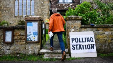 A man walking into a polling station in a rural church . He is seen from behind walking through a gate and wearing an orange coat 