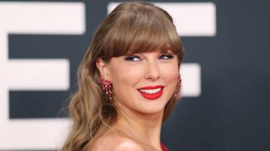 A photograph of Taylor Swift looking over her shoulder and smiling, wearing red earrings.