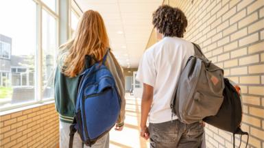 Two students walk along a corridor with their backs towards the camera. The student on the left has long red hair and a blue rucksack. The student on the right has shorter, brown hair and a grey rucksack.