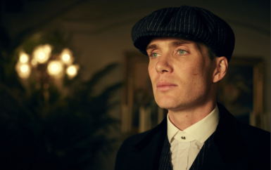 Cillian Murphy as his character Tommy in Peaky Blinders. He is dressed in formal, vintage-inspired attire. He wears a dark pinstripe flat cap, paired with a crisp white shirt fastened by a collar pin and layered beneath a dark coat. His light-colored eyes are striking, and a visible scar on his right chee.  The background is softly lit with warm tones, featuring blurred elements like a leafy plant and framed artwork. 