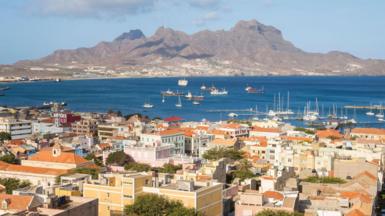 View over Mindelo, Sao Vicente, Cape Verde showing buildings with terracotta-coloured roofs in front of the blue ocean, which has several boats in it. Behind the ocean is a grey mountain and the sky is light and blue. 
