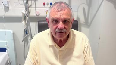 A man is sitting on a chair in a hospital ward. He has grey hair and a moustache. He is wearing a pale yellow polo top. Behind him is a hospital bed and numerous wires plugged into a plug socket on the wall. 