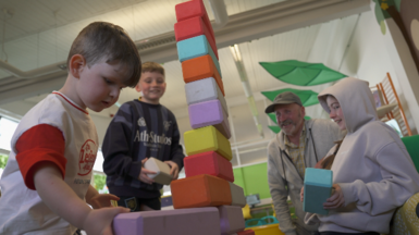A group of people in a play area watch a child stacking large foam blocks into a tall tower. Two other children and an adult sit nearby holding blocks. The setting includes bright colours, soft play equipment and decorative leaf shapes overhead.