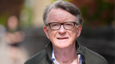 Mandelson pictured walking down a London street. He is dressed casually in a green raincoat and an open necked shirt. 